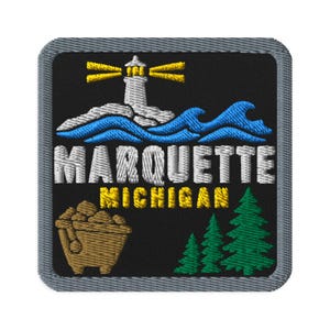 Puede incluir: Un parche bordado cuadrado con fondo negro y borde gris. Presenta un faro blanco con rayos de luz amarillos, olas azules, una vagoneta minera marrón y pinos verdes. El texto dice MARQUETTE en blanco y MICHIGAN en amarillo.