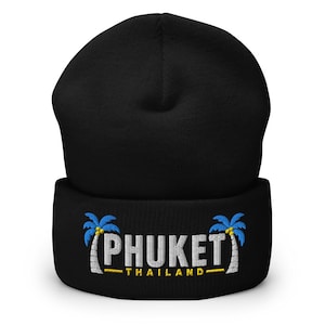 May include: A black beanie featuring an embroidered design on the cuff. The design includes two blue and white palm trees flanking the white text PHUKET, with the yellow text THAILAND positioned directly underneath the word PHUKET.