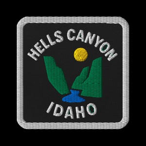 May include: Embroidered patch featuring the words "HELLS CANYON IDAHO" in white, arched over a landscape scene. The scene includes green mountains, a blue river, and a yellow sun against a black background, framed by a white border.