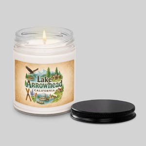 May include: A lit white candle in a clear glass jar with a black lid. The label features a vintage-style illustration with the text "Lake Arrowhead California" and an image of a lake, mountains, and buildings.