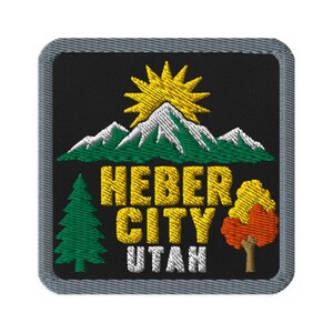 May include: A square embroidered patch with a black background and a gray border. It features a yellow sun rising over green and white mountains, a green pine tree, an orange and yellow deciduous tree, and the yellow text HEBER CITY UTAH.