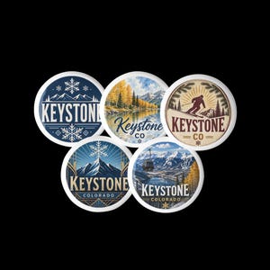 May include: Five circular pin-back buttons featuring various Keystone, Colorado designs. The graphics include mountain landscapes, a skier, a gondola, and snowflakes, with the text Keystone or Keystone CO printed on each individual button design.