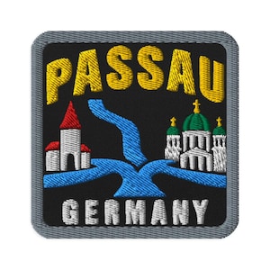 May include: A square embroidered patch with a black background and a grey border. The yellow text reads PASSAU at the top and white text reads GERMANY at the bottom. It features a blue river confluence, a white building with a red roof, and a white church.