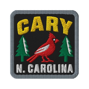 Puede incluir: Un parche bordado cuadrado con borde gris sobre fondo negro. El texto CARY aparece en amarillo en la parte superior y N. CAROLINA en blanco en la parte inferior. Un pájaro cardenal rojo está centrado entre dos pinos verdes con detalles blancos.