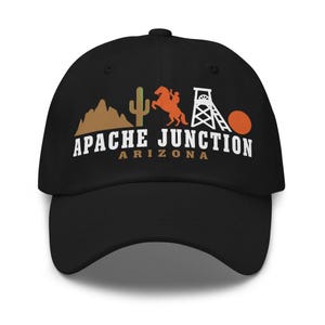 Op de afbeelding: Een zwarte baseballpet met een grafische print van een bruine berg, een groene cactus, een oranje paard met ruiter, een witte mijnbouwstructuur en een oranje cirkel. De tekst op de pet luidt APACHE JUNCTION ARIZONA in witte letters.
