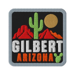 Könnte beinhalten: Ein quadratischer Stickaufnäher mit schwarzem Hintergrund, der einen grünen Saguarokaktus, rote Wüstenberge und eine beigefarbene Sonne zeigt. Der weiße Schriftzug lautet GILBERT und der orangefarbene ARIZONA, mit einem kleinen grünen Kaktus unten rechts.