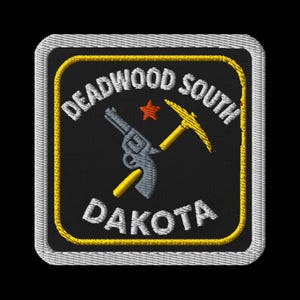Könnte beinhalten: Gestickter Aufnäher mit den Worten "Deadwood South Dakota" in Weiß, einer silbernen Pistole, einer gelben Spitzhacke und einem roten Stern auf schwarzem Hintergrund, umrahmt von einem gelben Rand und einer weißen Naht.