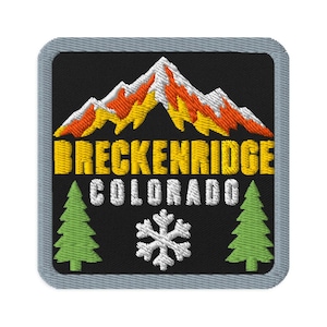 May include: A square embroidered patch with a gray border on a black background. It features a mountain range with white, yellow, and orange peaks above the yellow text BRECKENRIDGE and white text COLORADO, flanked by two green pine trees and a snowflake.