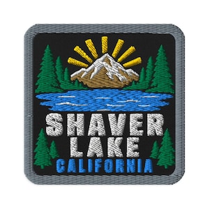 May include: A square embroidered patch with a black background featuring a brown mountain with white snow, a blue lake, green pine trees, and yellow sun rays. The text SHAVER LAKE CALIFORNIA is embroidered in white and blue block lettering.