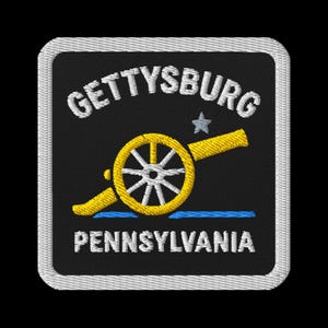 Puede incluir: Parche bordado con el texto "GETTYSBURG PENNSYLVANIA" en blanco. Un cañón amarillo con una rueda blanca y una línea azul están en el centro. El parche tiene un fondo negro y un borde blanco.