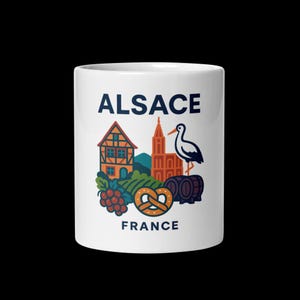 May include: White ceramic mug featuring a colorful illustration of Alsace, France. The design includes a traditional house, church, stork, barrels, grapes, and a pretzel. The word "ALSACE" is printed above "FRANCE".