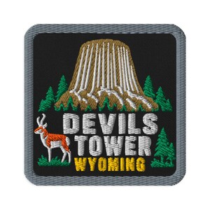 May include: A square embroidered patch with a black background and a gray border. It features a brown and white depiction of Devils Tower, green pine trees, a white and orange pronghorn, and the text DEVILS TOWER WYOMING in white and yellow.
