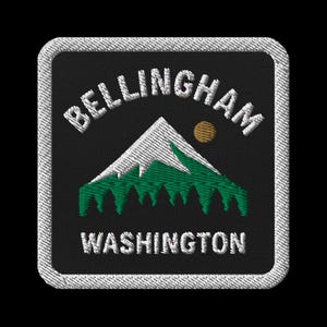May include: Embroidered patch with the text "BELLINGHAM WASHINGTON" in white, arched above a mountain scene. The mountain has a white peak and green base, with a yellow sun. The patch has a black background and a white stitched border.