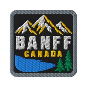 Könnte beinhalten: Ein quadratischer gestickter Aufnäher mit schwarzem Hintergrund und grauem Rand. Er zeigt weiße und gelbe Berggipfel über dem weißen Text BANFF und dem gelben Text CANADA. Ein blauer See und zwei grüne Tannen befinden sich unten.