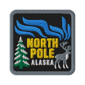 Könnte beinhalten: Ein quadratischer Stickaufnäher mit schwarzem Hintergrund und grauem Rand. Er zeigt einen grünen Tannenbaum mit weißem Schnee, ein graues Rentier und blaue Wellenlinien, die Nordlichter darstellen. Der Text NORTH POLE ALASKA ist gelb.