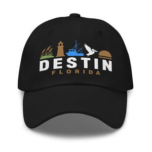 May include: A black baseball cap featuring a graphic design with a green plant, a brown lighthouse, a blue fishing boat, a white pelican, and a brown sun. The text DESTIN is printed in large white letters with FLORIDA in smaller brown letters below.