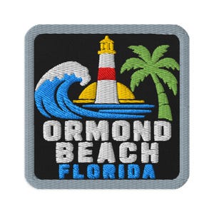 Puede incluir: Un parche bordado cuadrado con fondo negro y borde gris. Presenta un faro blanco y rojo, un sol amarillo, una ola azul y una palmera verde. El texto dice ORMOND BEACH FLORIDA en letras de color blanco y azul.