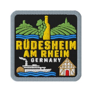 Puede incluir: Un parche bordado cuadrado con fondo negro y borde gris. Presenta una botella de vino amarilla, uvas verdes, colinas verdes, agua azul, un barco fluvial blanco y una casa de entramado de madera. El texto dice RÜDESHEIM AM RHEIN GERMANY.