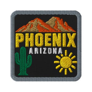 Könnte beinhalten: Ein quadratischer gestickter Aufnäher mit grauem Rand auf schwarzem Hintergrund. Er zeigt orangefarbene und beige Berge über dem gelben Schriftzug PHOENIX und dem weißen Schriftzug ARIZONA. Ein grüner Saguaro-Kaktus ist links und eine gelbe Sonne rechts.