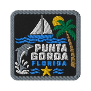 Puede incluir: Un parche bordado cuadrado con fondo negro y borde gris. Presenta un velero blanco sobre olas azules, un sol amarillo, una palmera verde, un delfín gris saltando sobre agua blanca, una estrella de mar amarilla y el texto PUNTA GORDA FLORIDA.