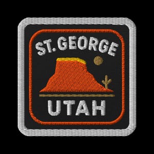 Puede incluir: Parche bordado con "ST. GEORGE UTAH" en letras blancas. El diseño incluye una montaña roja y amarilla, un sol y un cactus sobre un fondo negro, con un borde naranja y un borde blanco cosido.