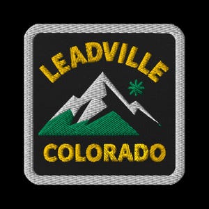 以下が含まれることがあります： 「LEADVILLE COLORADO」の文字が黄色で刺繍されたパッチ。山脈のグラフィックが上下に配置されています。山は白と緑で、小さな緑色の星があります。パッチは黒い背景と白い縁取りが施されています。