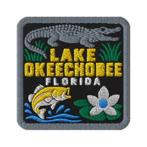 May include: A square embroidered patch with a black background and a grey border. It features a grey alligator, green grass, a yellow fish jumping in blue water, a white flower on green leaves, and the yellow text LAKE OKEECHOBEE and white FLORIDA.