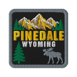 Può includere: Una toppa ricamata quadrata con sfondo nero raffigurante una catena montuosa con cime dorate e bianche, pini verdi e la sagoma di un alce grigio. Il testo PINEDALE è in giallo e WYOMING in bianco, il tutto bordato da un bordo grigio.
