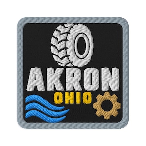 Pode incluir: Um emblema bordado quadrado com fundo preto e borda cinza. Apresenta um gráfico de pneu branco acima do texto branco AKRON, com o texto amarelo OHIO abaixo, acompanhado por linhas onduladas azuis e um ícone de engrenagem dourado.