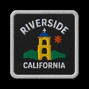 Puede incluir: Parche bordado con la palabra "RIVERSIDE" sobre una torre amarilla con cúpula azul y cruz, y "CALIFORNIA" debajo. El diseño incluye un sol naranja y colinas verdes sobre un fondo negro.
