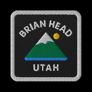 May include: Embroidered patch featuring the words "BRIAN HEAD UTAH" in white arched text above a mountain scene. The mountain has a white peak, green slopes, and a blue base, with a yellow sun. The patch has a black background and a white border.