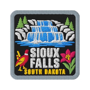 Op de afbeelding: Een vierkante geborduurde patch met een grijze rand op een zwarte achtergrond. Het toont een blauwe waterval, groene dennenbomen, een rode en gele fazant, een roze bloem en de witte tekst SIOUX FALLS boven gele tekst SOUTH DAKOTA.