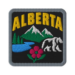 Puede incluir: Un parche bordado cuadrado con fondo negro y borde gris. El texto ALBERTA está bordado en amarillo en la parte superior. Debajo, una cordillera verde con picos blancos, un lago azul, la silueta de un oso blanco y una flor rosa.