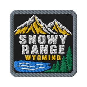May include: A square embroidered patch with a gray border on a black background. It features white and yellow mountain peaks above the white text SNOWY RANGE and yellow text WYOMING, with a blue lake and green pine trees at the bottom.