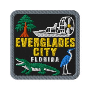 Könnte beinhalten: Ein quadratischer Stickaufnäher mit schwarzem Hintergrund und grauem Rand. Er zeigt eine grüne Zypresse, ein weißes Sumpfboot, einen grünen Alligator und einen blauen Reiher. Der Text lautet EVERGLADES CITY in Gelb und FLORIDA in Weiß.