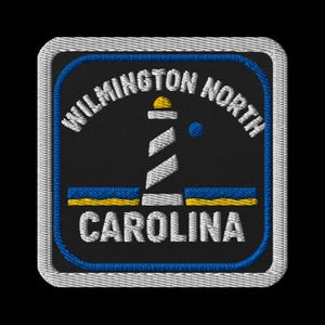 以下が含まれることがあります： 灯台のデザインと「WILMINGTON NORTH CAROLINA」の文字が刺繍されたパッチ。パッチは黒い背景に白、青、黄色のアクセントがあります。灯台は白で、上部は黄色です。