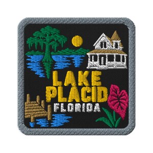 Puede incluir: Un parche bordado cuadrado con fondo negro y borde gris. Presenta un ciprés verde, un sol amarillo, una casa blanca, un muelle de madera y una flor rosa. El texto dice LAKE PLACID FLORIDA en letras de color amarillo y blanco.