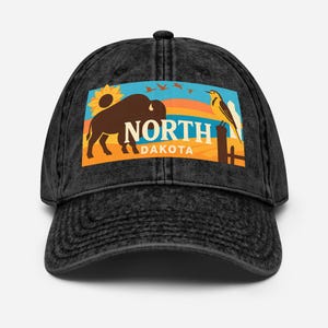May include: Black baseball cap with a faded appearance. The front displays a rectangular graphic with a bison, sunflower, and bird, and the words "NORTH DAKOTA" in white. The background has a sunset design.