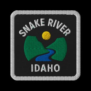 May include: Embroidered patch with the text "SNAKE RIVER IDAHO" in white, above a landscape scene. The scene shows a yellow sun, green mountains, and a blue river. The patch has a black background and a white border.