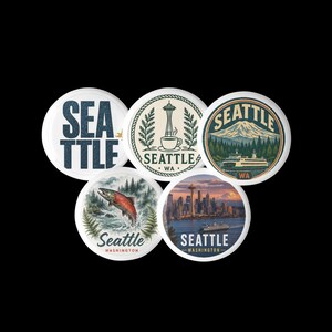 May include: A set of five circular buttons featuring Seattle-themed designs. One displays the text SEATTLE, another shows the Space Needle with a coffee cup, a third depicts a mountain and ferry, a fourth shows a salmon, and the fifth a city skyline.