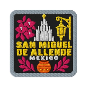 May include: A square embroidered patch with a black background and a gray border. It features a white church, a yellow street lamp, pink flowers, and an orange pot. The text reads SAN MIGUEL DE ALLENDE MEXICO in yellow and white lettering.
