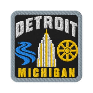 May include: A square embroidered patch with a black background and a grey border. The text DETROIT is in white at the top, and MICHIGAN is in yellow at the bottom. It features a white and yellow skyscraper, a blue river, and a yellow wheel.