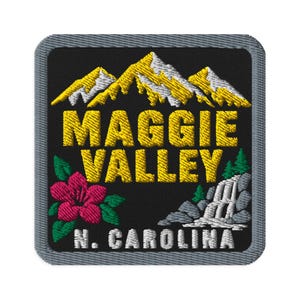 Può includere: Una toppa ricamata quadrata con sfondo nero e bordo grigio. Presenta cime montuose gialle e bianche, un fiore rosa con foglie verdi, una cascata su rocce grigie e il testo MAGGIE VALLEY N. CAROLINA in giallo e bianco.