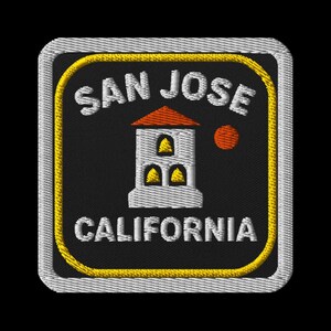 Op de afbeelding: Geborduurde patch met de tekst "SAN JOSE" en "CALIFORNIA". Het ontwerp toont een wit gebouw met een rood dak en een oranje cirkel. De patch heeft een zwarte achtergrond met een gele en witte rand.
