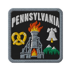 Puede incluir: Un parche bordado cuadrado con fondo negro y borde gris. Presenta el texto blanco PENNSYLVANIA en la parte superior, un pretzel amarillo, un horno industrial gris con llamas naranjas, una campana blanca y montañas verdes.