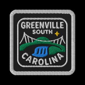 Op de afbeelding: Geborduurde patch met de tekst "GREENVILLE SOUTH CAROLINA" in wit, gebogen rond een centraal ontwerp. Het ontwerp toont een brug, groene heuvels, een blauwe waterval en een gele ster op een zwarte achtergrond, alles binnen een vierkant kader.