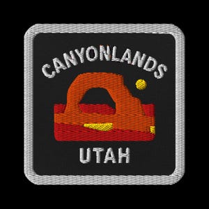 Pode incluir: Patch bordado com as palavras "Canyonlands Utah" em branco e um desenho de arco laranja e vermelho. O patch tem um fundo preto e uma borda branca.