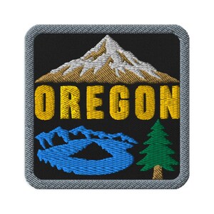 May include: A square embroidered patch with a black background and a gray border. It features a tan and white mountain peak above the yellow text OREGON. Below the text is a blue lake with a green pine tree on the right side of the patch.
