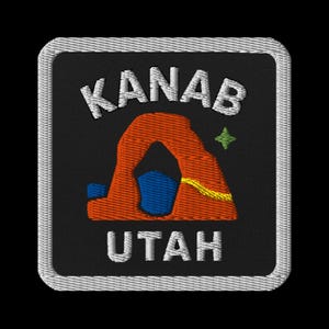 Pode incluir: Patch bordado com a palavra "KANAB" acima de um arco laranja e "UTAH" abaixo. O patch tem um fundo preto com uma borda prateada e uma estrela verde. O arco tem detalhes em azul e amarelo.