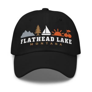 May include: A black baseball cap featuring a graphic design with a grey mountain, a brown pine tree, a white sailboat, an orange sun, and two orange cherries. Below the icons, the text reads FLATHEAD LAKE in white and MONTANA in brown.